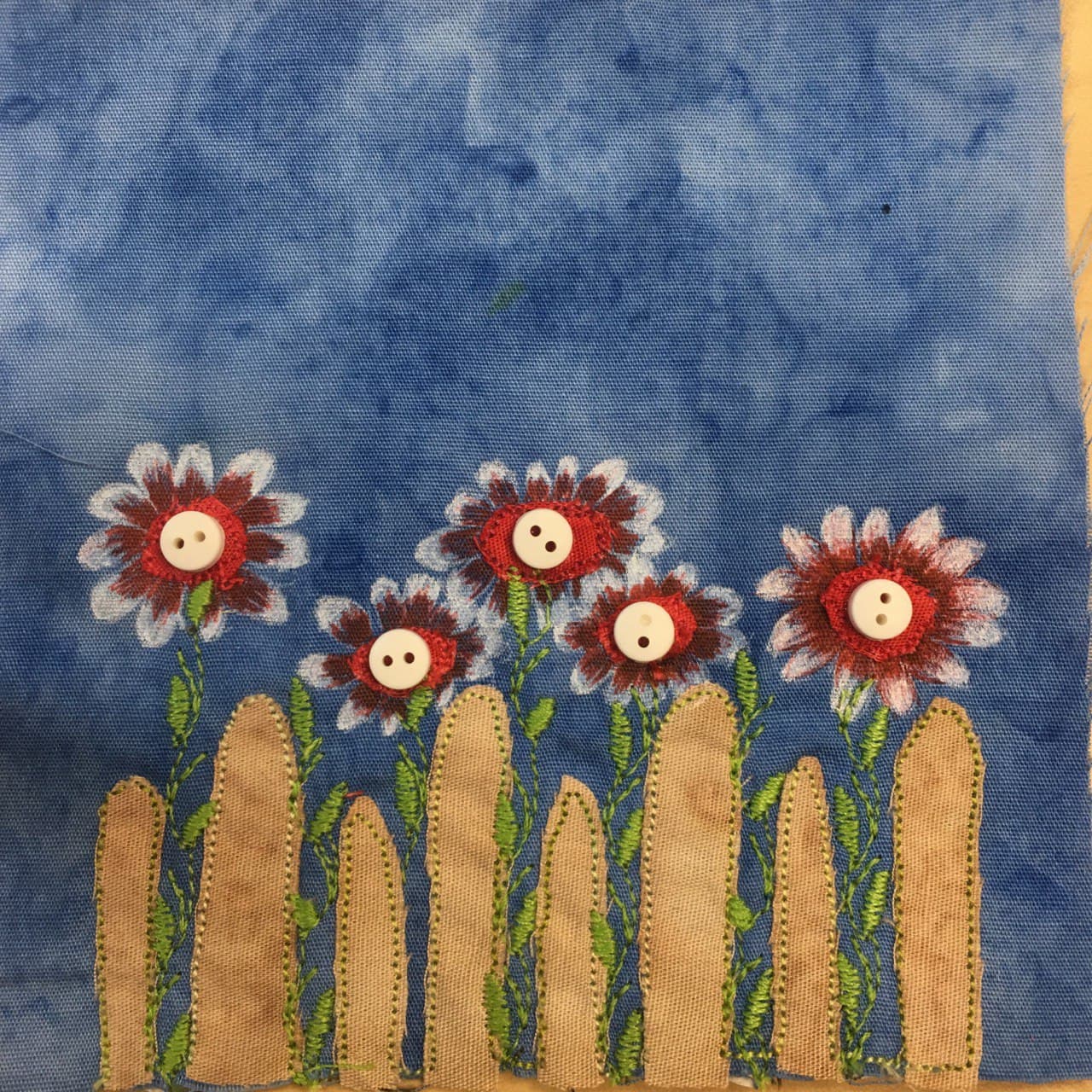 This quilt block shows a tan fence with slats that have significant gaps between them. The slats are rounded at the end. In between the slats and above them are a row of beautiful flowers. The greenery for the flowers is done with intricate stitching, with even the leaves as stitches. The flowers are a combination of red fabric with medium sized two-hole buttons in the center, and red and white pen marking coming out from the center to show beautiful flower petals. This is all against a blue salmon-patterned background.