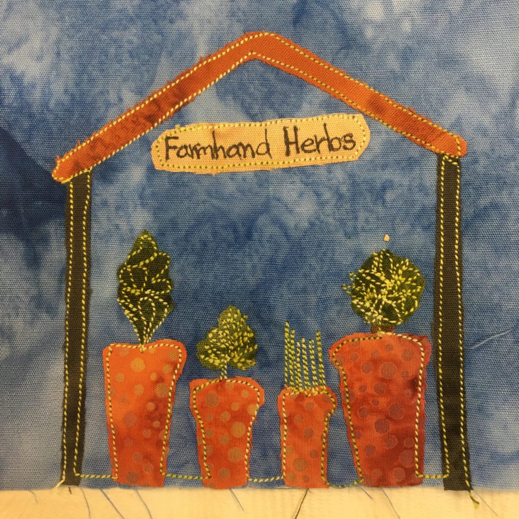 This quilt block shows a roadside herb stand. The stand is made up of two dark sticks on the side and a triangular thin ceiling that is orange. Inside the stand are four pots in a row, each a piece of orange polka-dotted duotone fabric. On the top of each pot is a piece of green fabric. Each piece is a different shape and heavily stitched to indicate its shape. The shack and pots are outlined with a bright yellow stitching. This is all against a blut salmon-patterned background.