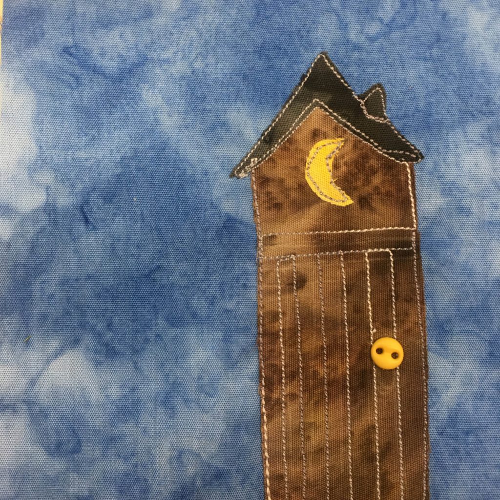 This quilt block shows an outhouse against a blue salmon-patterned background. The outhouse is a dark brown with white stitching on it to indicate the door and slats. The roof is almost black and is a thin triangular shape. On the front of the door is a small two-holed yellow button for the door handle. At the top of the outhouse is a yellow crescent moon.