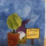 This quilt block shows a small young round tree, a sign and a purple hatted garden gnome all sitting next to each other. The tree has a green ball for it's greenery, and a orange-brown terracotta looking pot (that's really plastic). The garden gnome's eyes are obscured by his hat, and his beard is white. His little nose is a pink color. The nose and beard are marked with pen. He's wearing a green shirt with a floral design printed on it. Next to him is a yellow sign that reads in part cursive, 