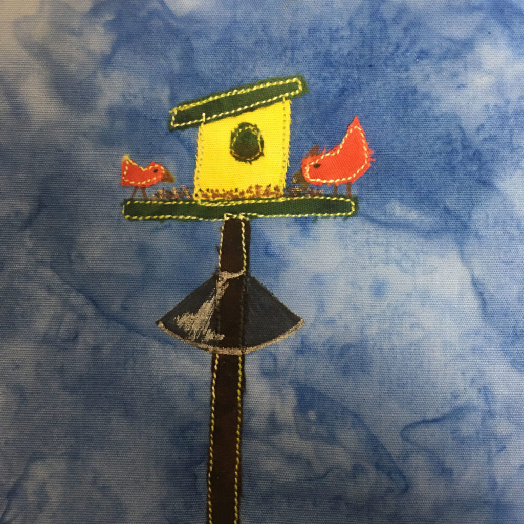 This quilt block shows a bird feeder with two birds on it against a blue salmon-patterned background. The bird feeder is on a black pole with yellow stitching outlining it. On the pole is a cone shape with the broad base at the bottom to keep out squirrels. The cone shape is drawn with pen and has a sheen on it. The bird feeder is yellow with a slanted green roof. The birds sitting on the perch are a red-orange with a yellow stitching outline and drawn beaks.