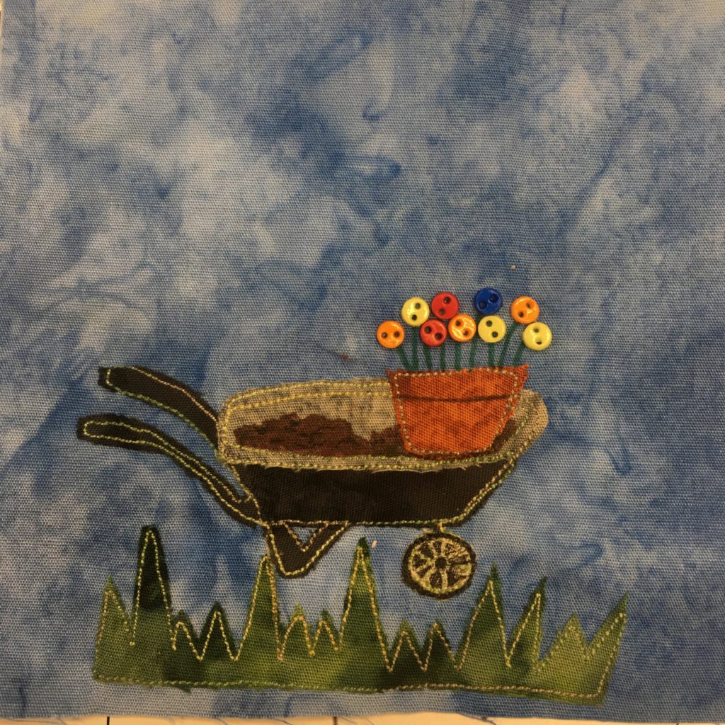 This quilt block shows a jagged piece of green fabric at the bottom to indicate grass. Above that strip of grass is a pretty detailed wheelbarrow constructed out of very dark brown fabric with a yellow stitching outline. The top rim and inside of the wheelbarrow is a gray fabric. Inside the wheelbarrow, which we are seeing from the side, is dirt, brown fabric marked with pen, and a flower pot. Inside the flower pot are pen lines going up into buttons. The flower bulbs are tiny two-hole buttons of various colors.
