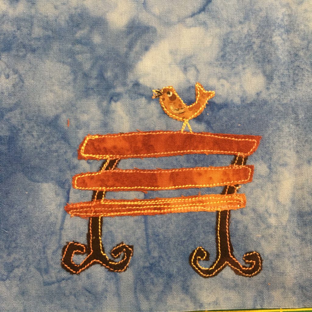This quilt block shows a golden bench against a blue salmon-patterned background. The bench's slats are made out of a golden orange fabric. The legs are curved to the right, and each ends in two curved feet that curl upwards towards the seat of the bench. The legs are much darker brown than the bench in a very complementary color. The whole bench is outlined with a bright yellow and orange fabric stitching, giving it a sort of glow. Perched on top of the bench is a yellow bird with a drawn beak and eyes in pen. It appears to be chirping.