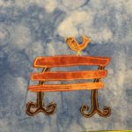 This quilt block shows a golden bench against a blue salmon-patterned background. The bench's slats are made out of a golden orange fabric. The legs are curved to the right, and each ends in two curved feet that curl upwards towards the seat of the bench. The legs are much darker brown than the bench in a very complementary color. The whole bench is outlined with a bright yellow and orange fabric stitching, giving it a sort of glow. Perched on top of the bench is a yellow bird with a drawn beak and eyes in pen. It appears to be chirping.