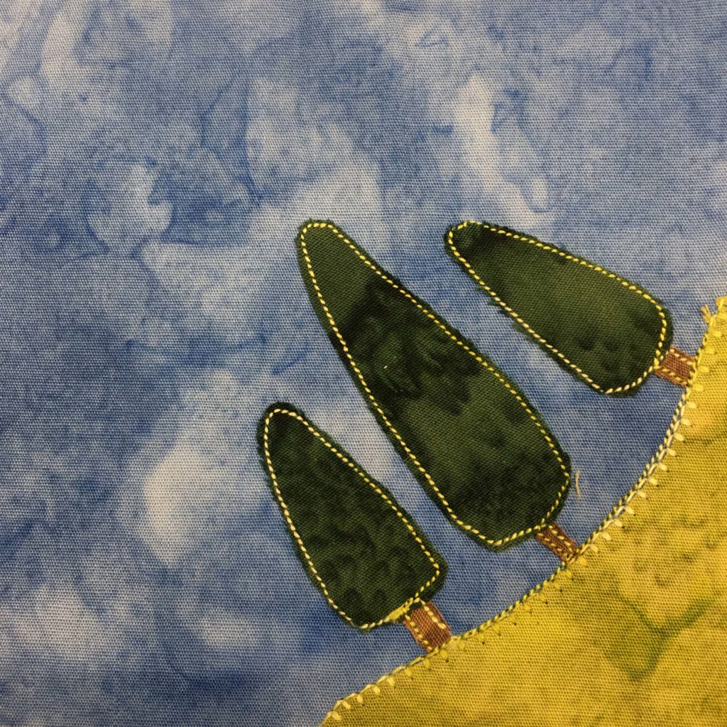 This quilt block shows a corner of well-tended grass in the bottom right. The grass is indicated by a piece of light green fabric with a strong big stitching on the edge of it. On top of the grass are three big dark green trees in cone shapes. The trunks are brown, but both the foliage and trunks are outlined by a similar yellow-green stitching. This is all against a blue salmon-patterned background.