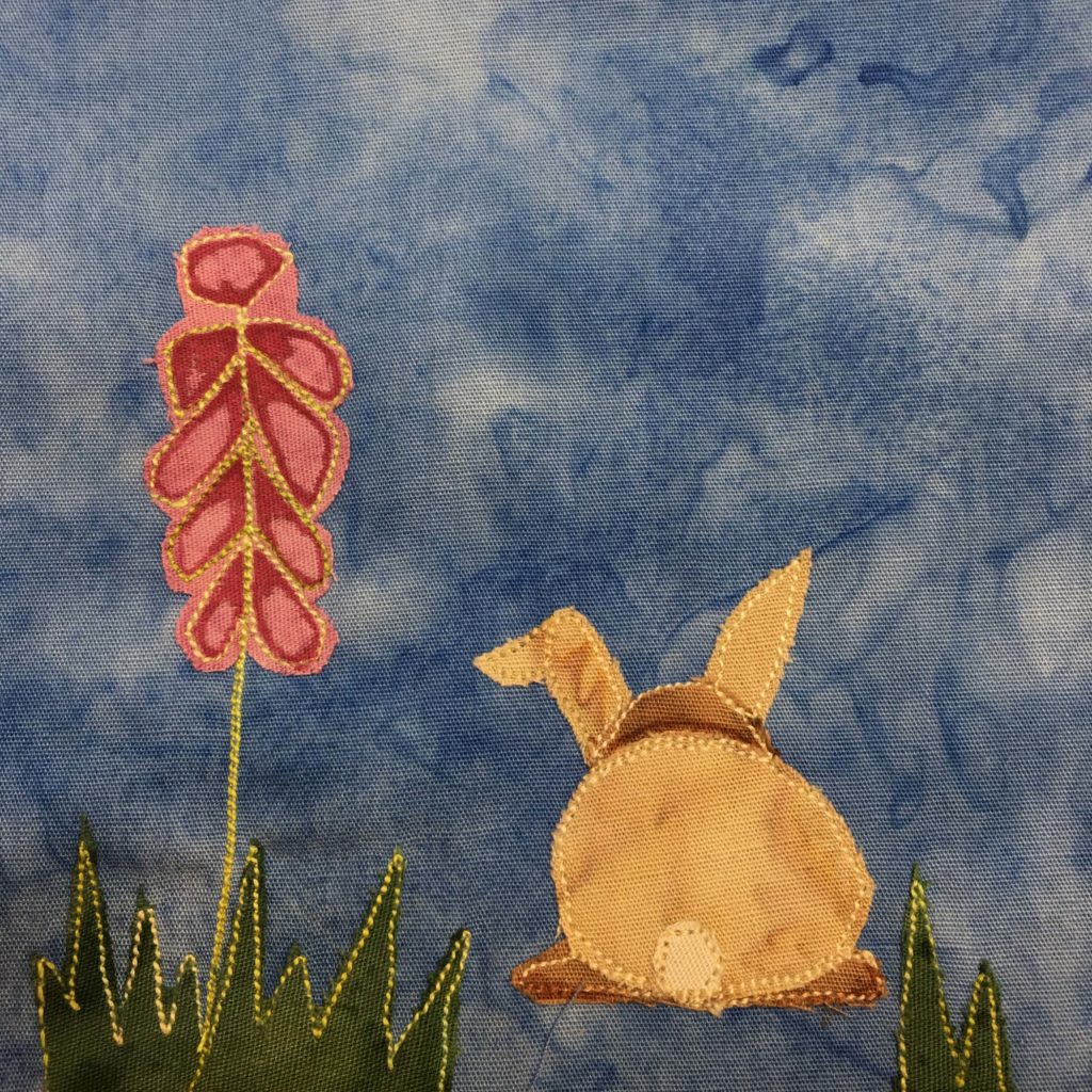 This quilt block shows two patches of grass, one big one on the bottom left and a small one on the bottom right. The grass patches are jagged cut green fabric outlined in yellow stithing. Coming out of the left patch of grass is a flower spike, a column of bulbs, that looks a bit like Gladiolus. The bulbs are colored pink and red (red with pen and pink fabric). Next to the spike of bulbs is a bunny viewed from the back. The bunny is tan, has a lighter tail, and two ears, one floppy.