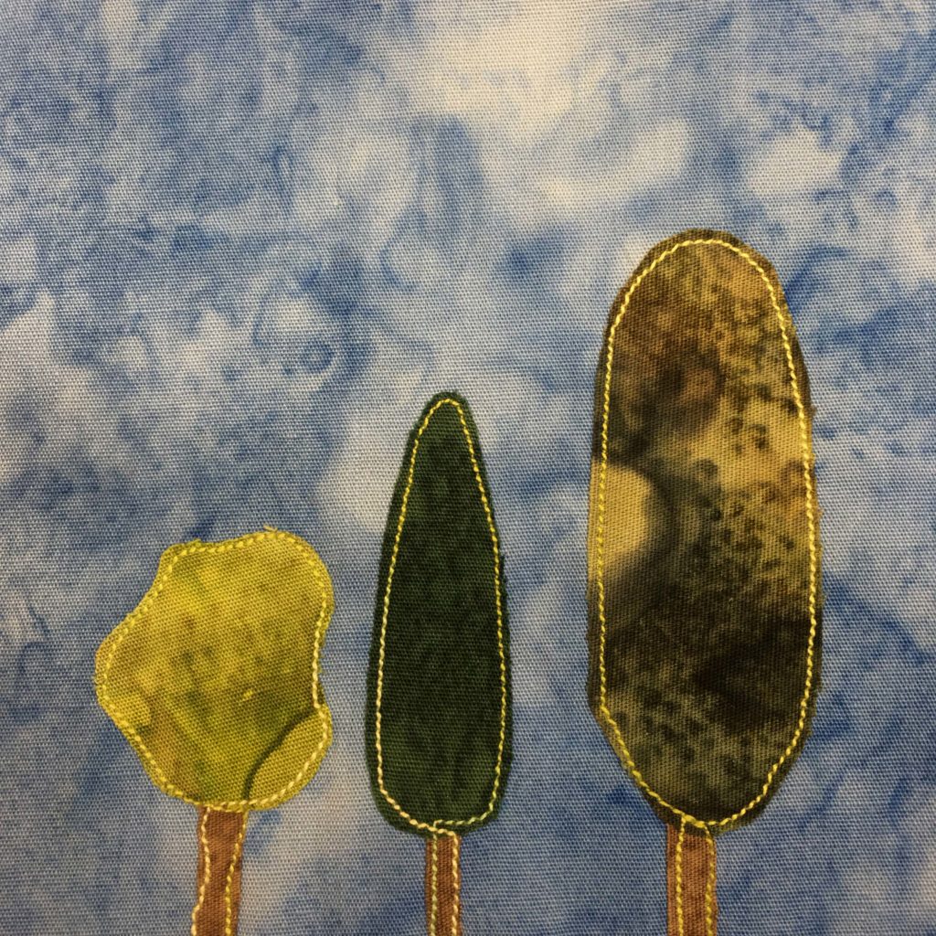 This quilt block shows three trees next to each other. Each tree is a different shape. The rightmost tree is tall and an oval shape and is a dark splotchy green. The tree to it's left is pretty standard, cone shaped, and is an even dark green. The tree farthest to the left is amorphous in shape, and is a bright green fabric. The trunks are all a light brown and outlined by a yellow stitching. This is all against a blue salmon-patterned background.