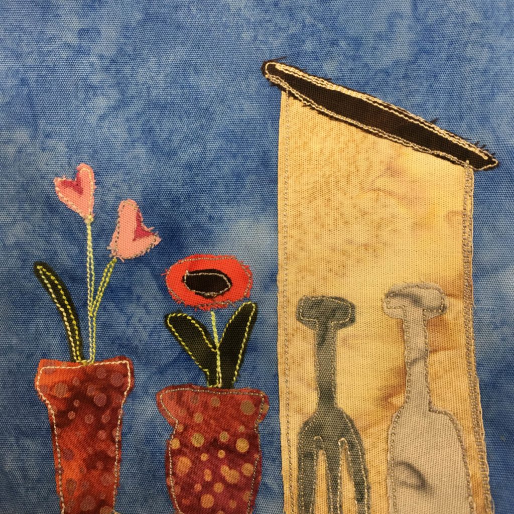 This quilt block shows a shed on the left side, and some tools on the side (front to us) of the shed. There is a gray pitchfork, and a light gray shovel. The shed itself is a piece of light tan orange fabric, with a dark brown roof that has yellow stitched outline. On the left side of the block are two potted flowers. The pots are a dark red with lighter duotone polka dots. The two flowers are different, one having two bulbs that are pink and red, and one being sunflower-like with a red outside and a black center.