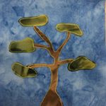 This quilt block shows a very large tree that spans the whole block. Its trunk is brown and branches off without foliage. Each branch has a green piece of fabric at the end of it indicating the location of the foliage. Each fabric piece is outlined with yellow stitching. This is against a blue salmon-patterned background.