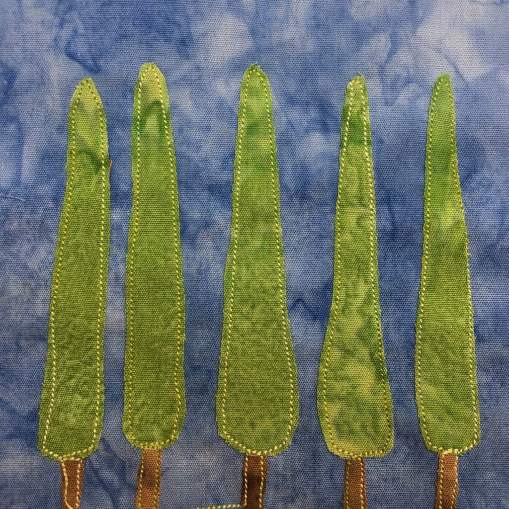 This quilt block shows five very tall pointy and skinny trees in a row against a blue salmon-patterned background. Each tree has a short little trunk at the bottom that is brown fabric, and then a long skinny and pointy foliage going up. The trunk and the greenery are outlined with yellow stitching.