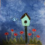 This quilt block shows a birdhouse with a bird against a blue salmon-patterned background. The birdhouse is near the center of the block and stands on a black pole. The house itself is a light blue with a black hole in the middle. The roof is angled like a triangle and is black. There is a patch of grass underneath indicated by a jagged cut piece of green fabric. Coming out of the grass are red flowers with long stems. Each flower is a red circle with a black middle. To the left of the birdhouse in the air is a bluebird. The bluebird is drawn in with pen and is in mid flight.