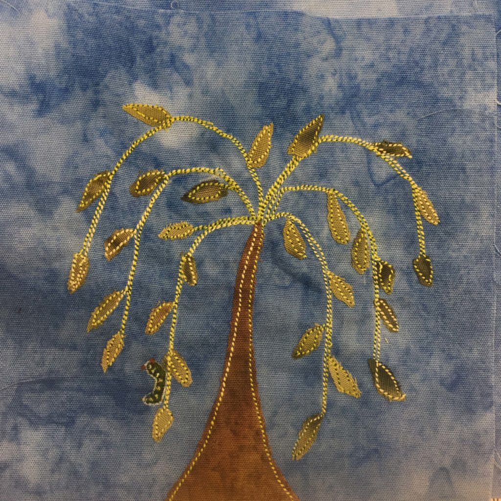 This quilt block shows a weeping willow tree. The trunk of the tree rests in the center of the bottom of the block and grows upward, tapering to the end point at the top. Then yellow and green stitching comes out of the top like a fountain and "sprays" downward. With each stitching line there are green leaves outlined by stitching to indicate the leaves of the tree. On the left leave vine closest to the trunk there is drawn a dark green caterpillar with yellow spots.