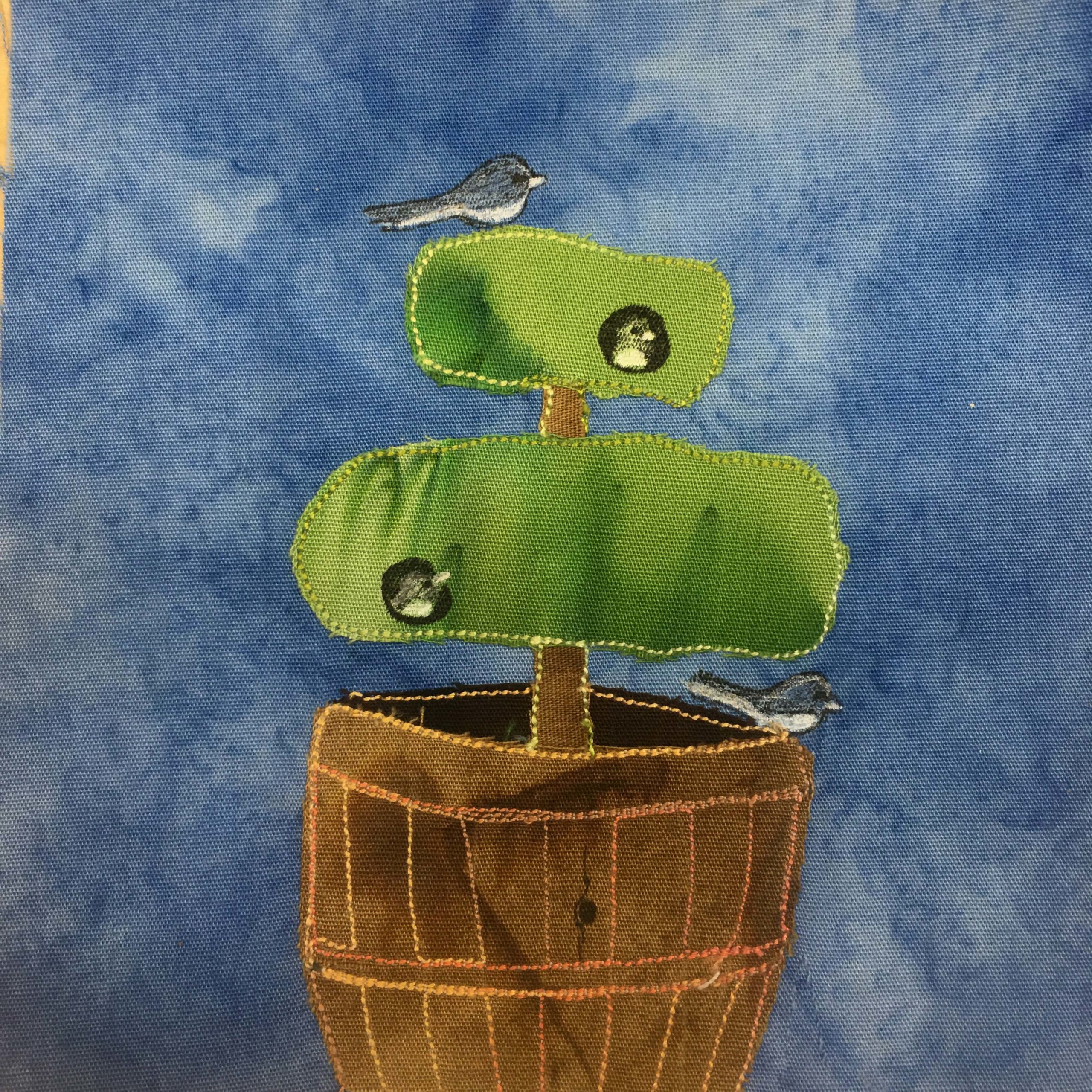 This quilt block shows a barrel made from brown boards with a dense topiary coming out of it. The topiary has been fashioned into two separate layers, with the lower layer being much larger. In this topiary are two bird heads poking out and looking around, one in the lower bush, and one in the upper bush. There is a small gray-white bird below the large layer on the barrel and a similar bird (a Junco bird) on the top topiary perching. The topiary is made of a green fabric. This is all against a blue background.