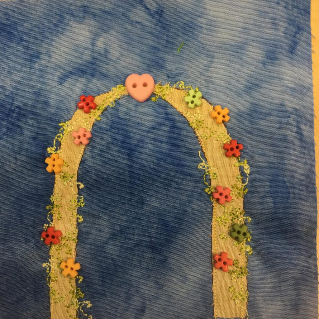 This quilt block shows an archway against a blue salmon-patterned background. The archway is one you would walk under for a wedding. It is covered with vines indicated by careful stitching, and flowers indicated by star shaped two holed buttons. The buttons are various colors ranging from orange to green. At the top of the archway is a larger pink heart button.
