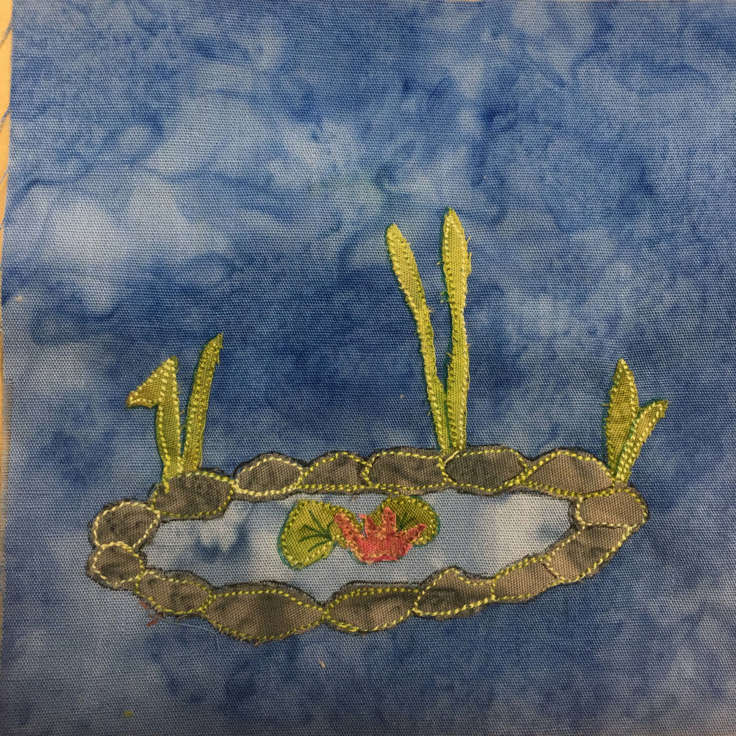 This quilt block shows a little garden water feature (pond) against a blue background. The pond is surrounded by gray rock made of gray fabric. The rocks are individually outlined in yellow stitching. There are three patches of tall grass leaves along the back side of the rocks made up of green fabric. And inside the pond, are a couple of lily pads and a red water flower, made up of green and red fabric with some extra pen drawn details. This is all against a blue salmon-patterned background.