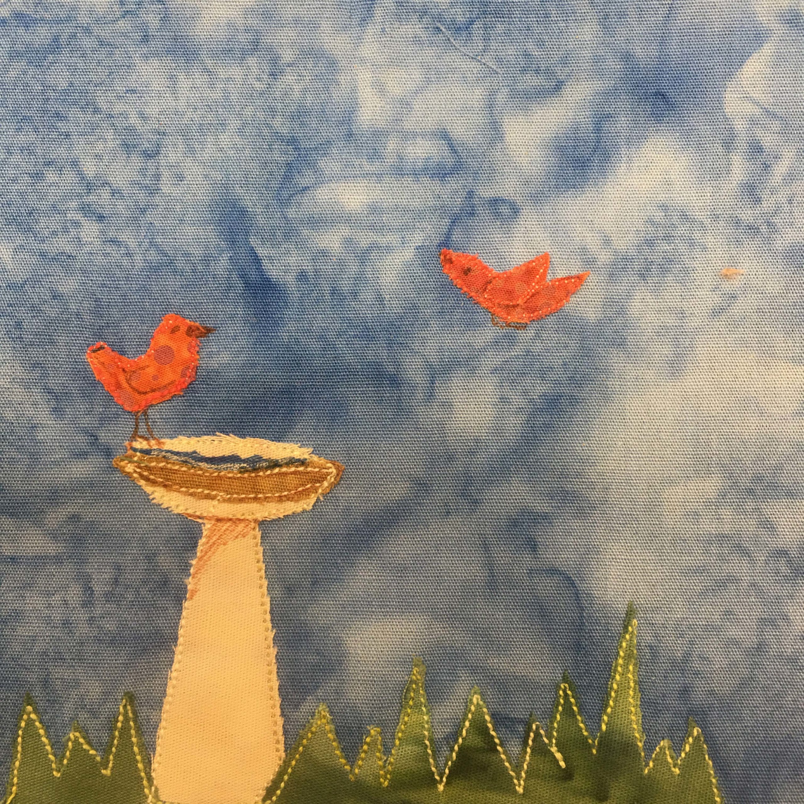 This quilt block shows a patch of grass at the bottom indicated by a dark green fabric cut with a jagged edge pointing up. The grass is outlined with a yellow stitching. To the left is a small birdbath. It's a simple tan cone with a small bowl at the top to make the bath. On top of the bowl, and about half as big as it, is a red bird. It is made up of duotone polka dotted red fabric. There is a similar bird mid-flight coming to rest on the other side of the birdbath. This is all against a blue salmon-patterned background.