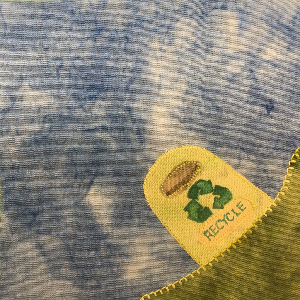 This quilt block shows a corner. In the bottom right is a green patch of grass. This grass is smooth and is bordered by a strong stitch. On top of the corner grass is a lime green recycle bin with a gray entry slot. The recycle bin has a dark green recycle symbol penned on it and below it is a sign that's barely yellow with the green word, "Recycle" written on it. This is against a blue salmon-patterned background.