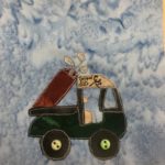 This quilt block shows a golf cart from the side facing right. It has two big dark wheels with a light off-white button on the back one, and a light green button on the front axle. The chassis of the cart is a very dark green fabric, while the frame is a black similar to the wheels. On the back it is carrying a golf bag that is maroon, and it has golf clubs that are silver poking out of it. The seat inside the cab, and the roof are a tan color,a nd the roof has penned on it a logo of an elk, and the words, 