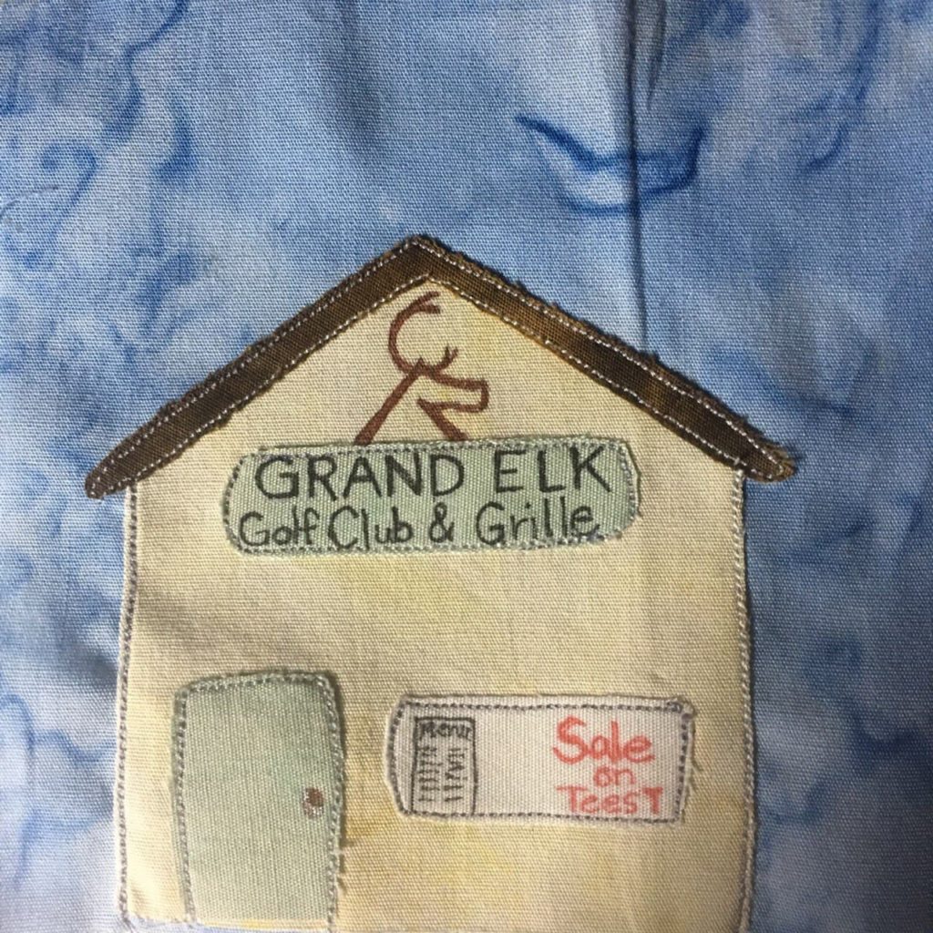 This quilt block shows a large square tan building against a blue salmon-patterned background. The building has a triangular sloped rooftop which is shown with a much darker brown fabric. Right at the apex of the roof is a logo of an elk. Underneath the elk logo is a sign that reads in black pen, "GRAND ELK Golf Club & Grille" The sign is in a gray or beige color. There is a window and inside the window is a menu pasted up, and in red pen, "Sale on Tees!" To the left of the window is a door the same color as the upper sign.