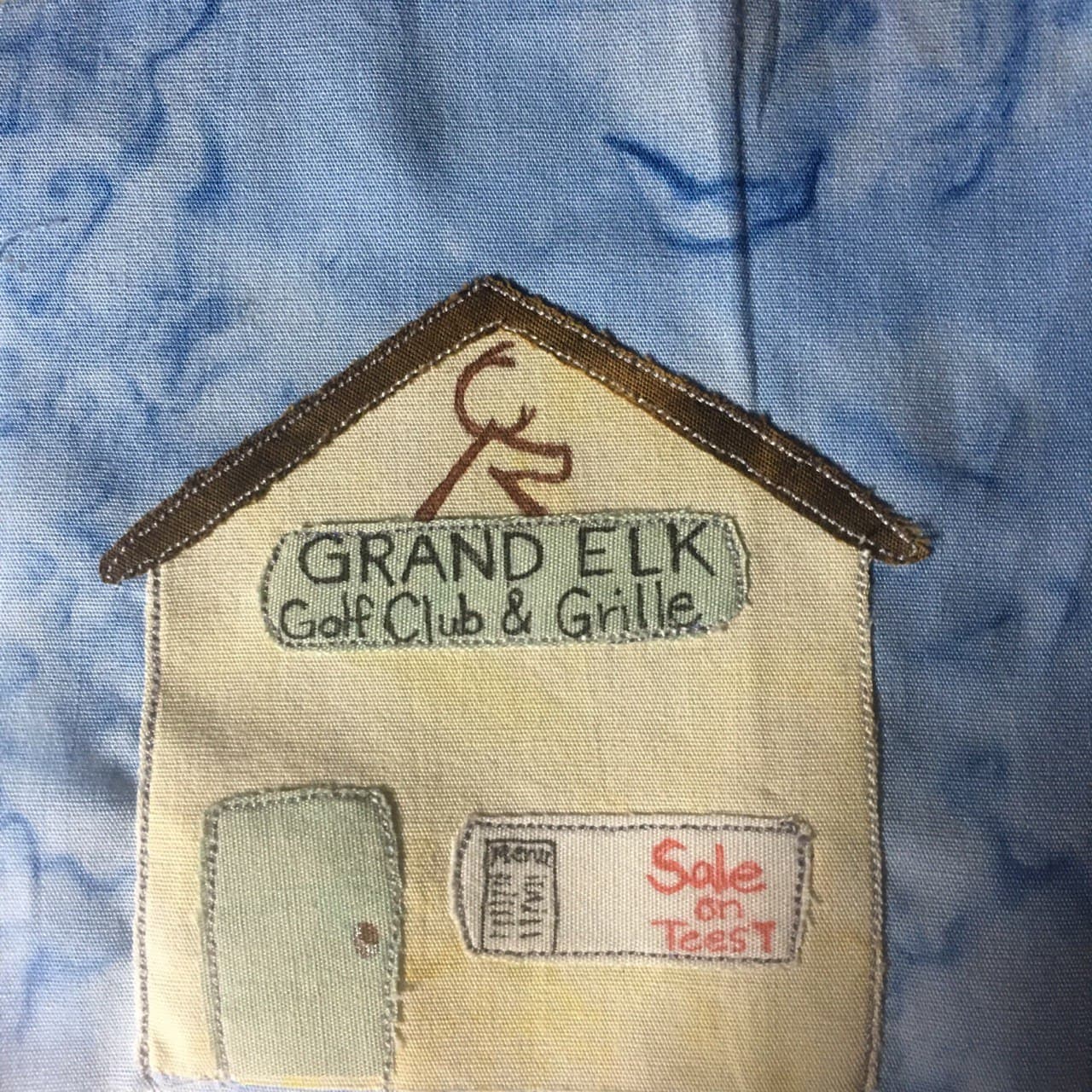 This quilt block shows a large square tan building against a blue salmon-patterned background. The building has a triangular sloped rooftop which is shown with a much darker brown fabric. Right at the apex of the roof is a logo of an elk. Underneath the elk logo is a sign that reads in black pen, "GRAND ELK Golf Club & Grille" The sign is in a gray or beige color. There is a window and inside the window is a menu pasted up, and in red pen, "Sale on Tees!" To the left of the window is a door the same color as the upper sign.