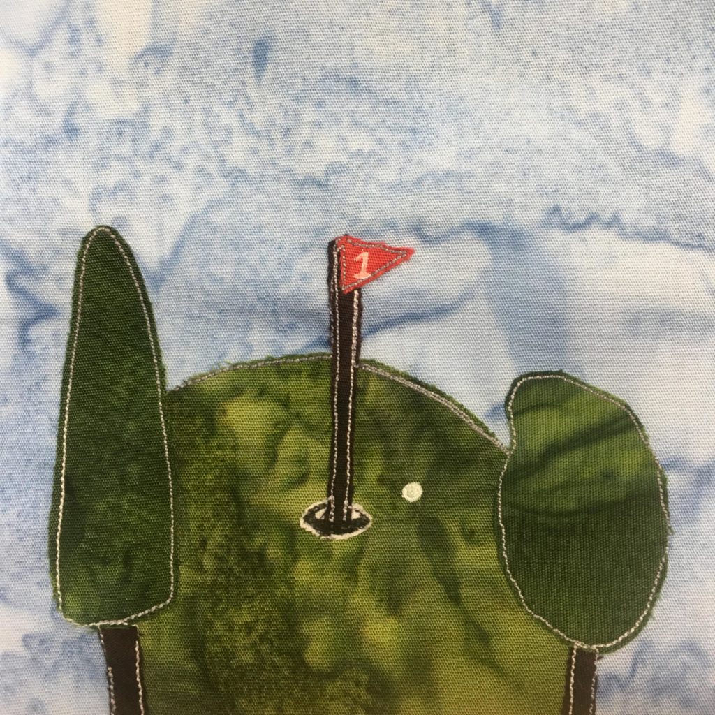 This quilt block shows a putting green on a golf course. There are two trees to either side of the putting green with black trunks and dark green fabric foliage. The putting green itself is the same dark green fabric, but in the center of the oval shaped surface is a big black pole sticking into a hole. At the top of the pole is a little red flag with the number 1 penned in white. Near the hole in the green is a white dot, also penned in. That's a golf ball,
Marc's golf ball.