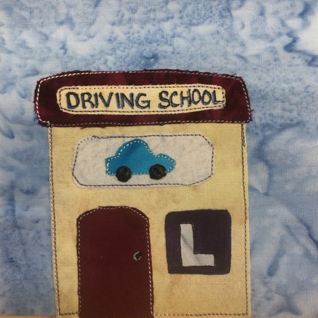 This quilt block shows a square building with a square roof. The building is a light tan, and the roof a very dark brown. On the roof is a light yellow sign with the words, "Driving School" written on it in pen. Below that is another sign, this one showing a blue car with white stitching outlining it. Below that is a dark brown door the same shade as the roof, and a large almost black sign with a gray upper case L in it. This is all against a blue salmon-patterned background.