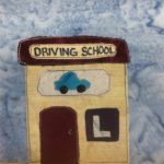 This quilt block shows a square building with a square roof. The building is a light tan, and the roof a very dark brown. On the roof is a light yellow sign with the words, "Driving School" written on it in pen. Below that is another sign, this one showing a blue car with white stitching outlining it. Below that is a dark brown door the same shade as the roof, and a large almost black sign with a gray upper case L in it. This is all against a blue salmon-patterned background.