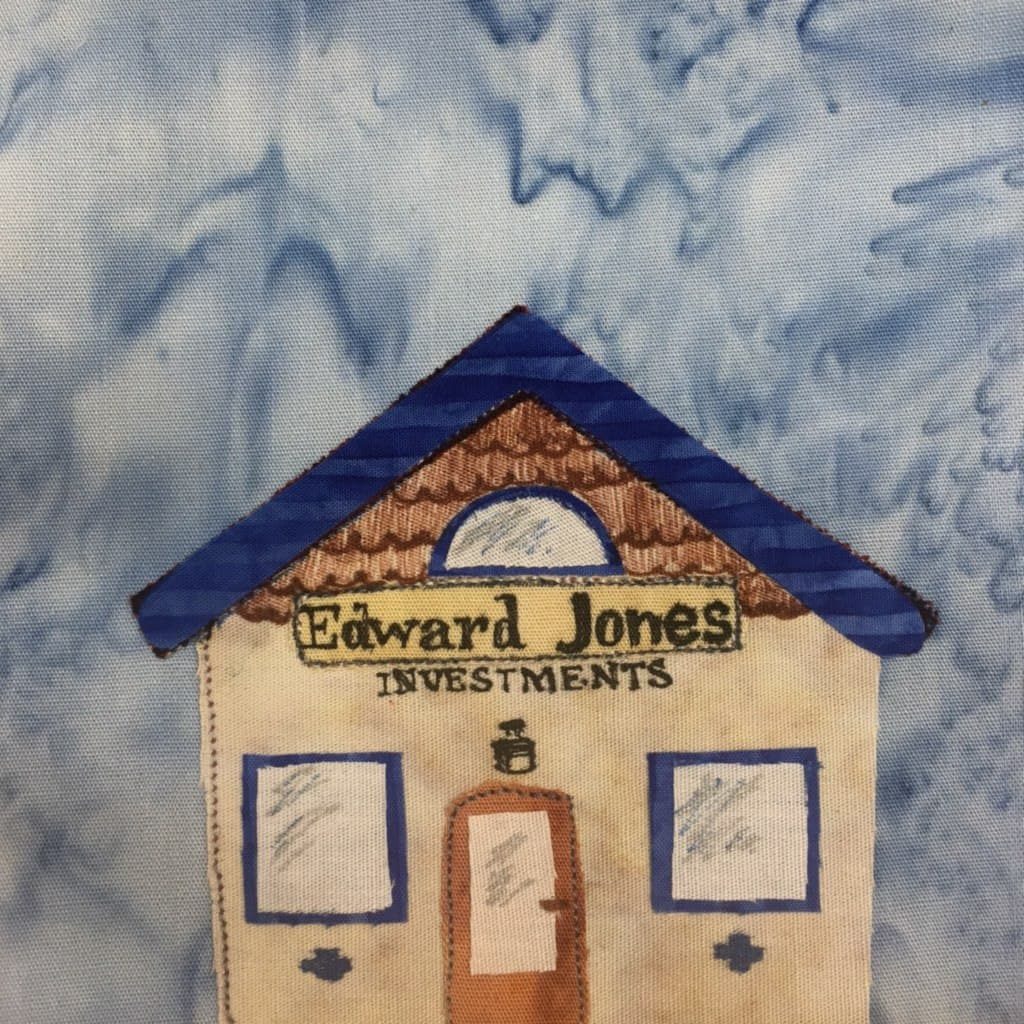 This quilt block features a short building with a triangular roof. The roof is a striped duotone deep blue with horizontal stripes. Below the roof is a half circle window surrounded by shingles that are brown and drawn in with a pen. Below the upper window is a sign in yello with the words "Edward Jones Investments" written in with pen. Below that is a light over the door. The door is brown but with a large glass pane inside it. On either side of the central door are blue trimmed windows. Each pane of glass is offwhite fabric with some sheen drawn in with pen to indicate it's reflective. Below the two blue windows are small blue crosses.