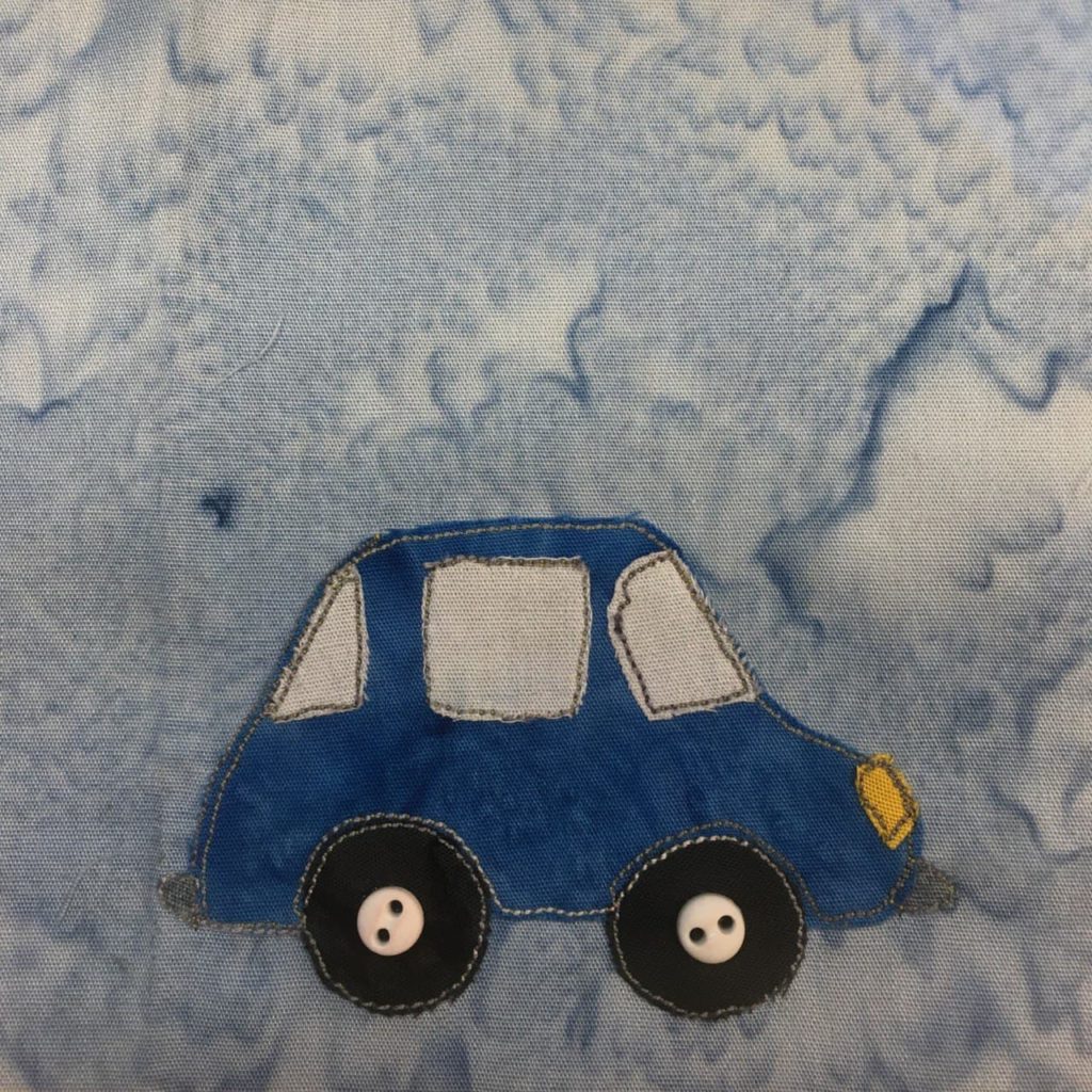 This quilt block shows a side view of a very nice car. The very nice car is a deep blue and has three windows: a windshield, a door window, and a rear window. There are two small gray bumpers on the front and back. It has two basic black circle wheels with two two-holed white small buttons in the center to indicate the axles. This is all against a light blue salmon-patterned background.