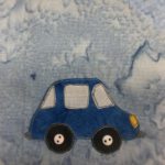 This quilt block shows a side view of a very nice car. The very nice car is a deep blue and has three windows: a windshield, a door window, and a rear window. There are two small gray bumpers on the front and back. It has two basic black circle wheels with two two-holed white small buttons in the center to indicate the axles. This is all against a light blue salmon-patterned background.