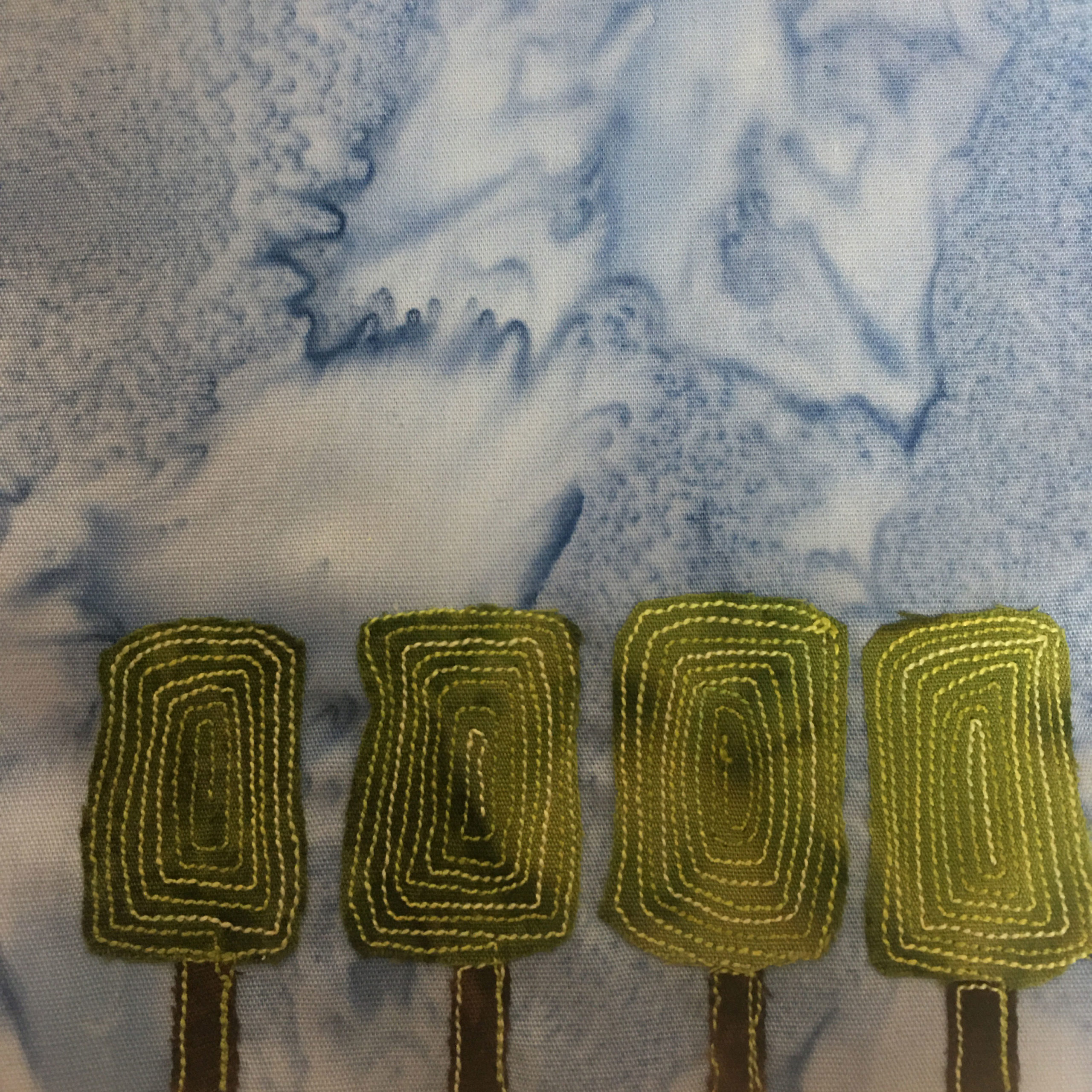 This quilt block shows four low shrubs all in a row at the bottom of the block. Each chrub is a square green topiary on top of a dark trunk. The trunk and topiary have light green stitching on them, the darker green topiary itself having a big swirl stitch on each. This is all against a light blue slamon-patterned background.