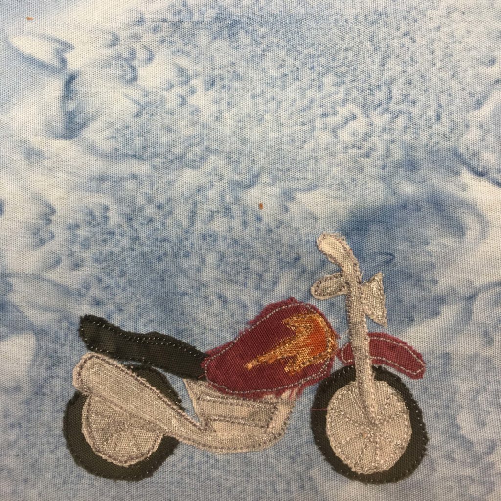 This quilt block shows a motorcycle made out of fabric. The chassis and motor are made out of a gray highly textured fabric and is accented with a light gray stitching indicating the engine and the tail pipe, as well as the handle bars. The wheels are a black circle of fabric, but mostly covered with the gray fabric indicating the spokes with the same stitching. The chrome is a dark red fabric with dark grey stitching, and on the main body behind the handlebars is an orange splotch of fabric in the shame o a flame. The seat is the same black fabric as the wheels, with black stitching.