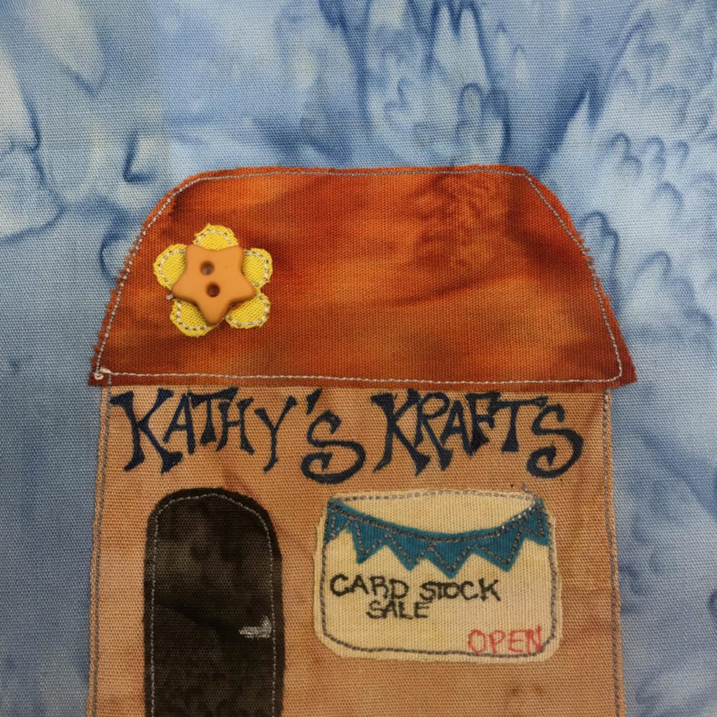 This quilt block shows a square building with a tall broad roof in the shape of a trapezoid (we're looking at it from the front). On the orange roof is a flower design, with a two-holed start button on a yellow piece of fabric. Underneath the burnt orange roof are the stylized words,