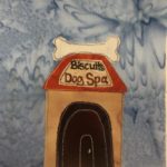 This quilt block shows a square building that is shaped like a doghouse. The door is a small door on the front center, but it's black to match the black of the large faux door painted on the front, as if Clifford the Big Red Dog lived there. On the orange roof is a yellow sign that reads,