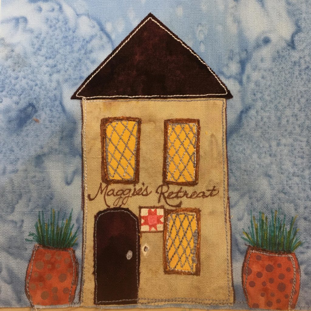 This quilt block shows a tall two story house. On either side of the house are two large plants with ferns coming out of them. The ferns are penned in green strokes, while the pots are duotone polka-dot fabrics. The house itself is a dark tan color with a very dark brown door on the left. To the right of the door is a quilt block on display in a frame, and to the right of that is a yellow window with a hatch pattern over it in gray. Above these items is writing in script that says,