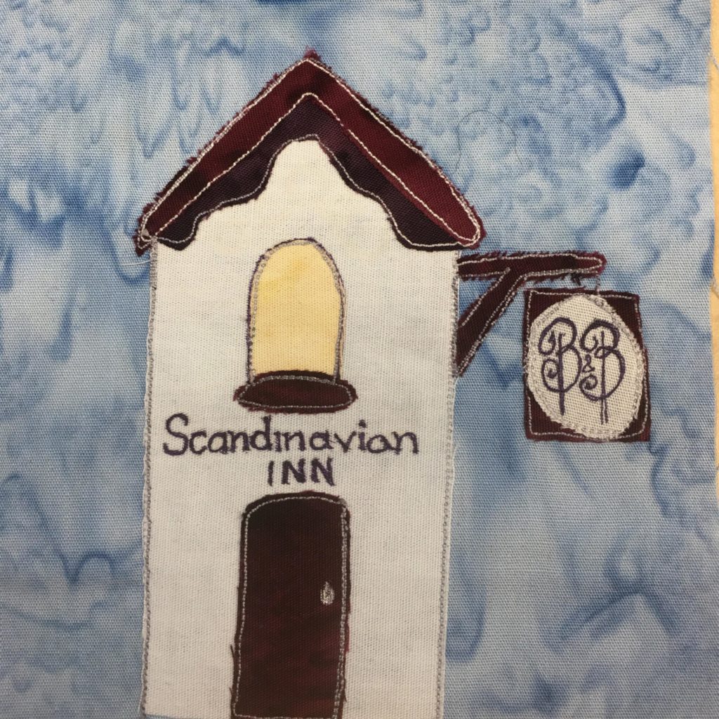 This quilt block shows a skinny tall white building with dark brown trimming. The roof is triangular with trimming underneath it. There is a yellow rounded window above the door with a window sill that is dark brown. Below that are the words, 