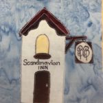 This quilt block shows a skinny tall white building with dark brown trimming. The roof is triangular with trimming underneath it. There is a yellow rounded window above the door with a window sill that is dark brown. Below that are the words,