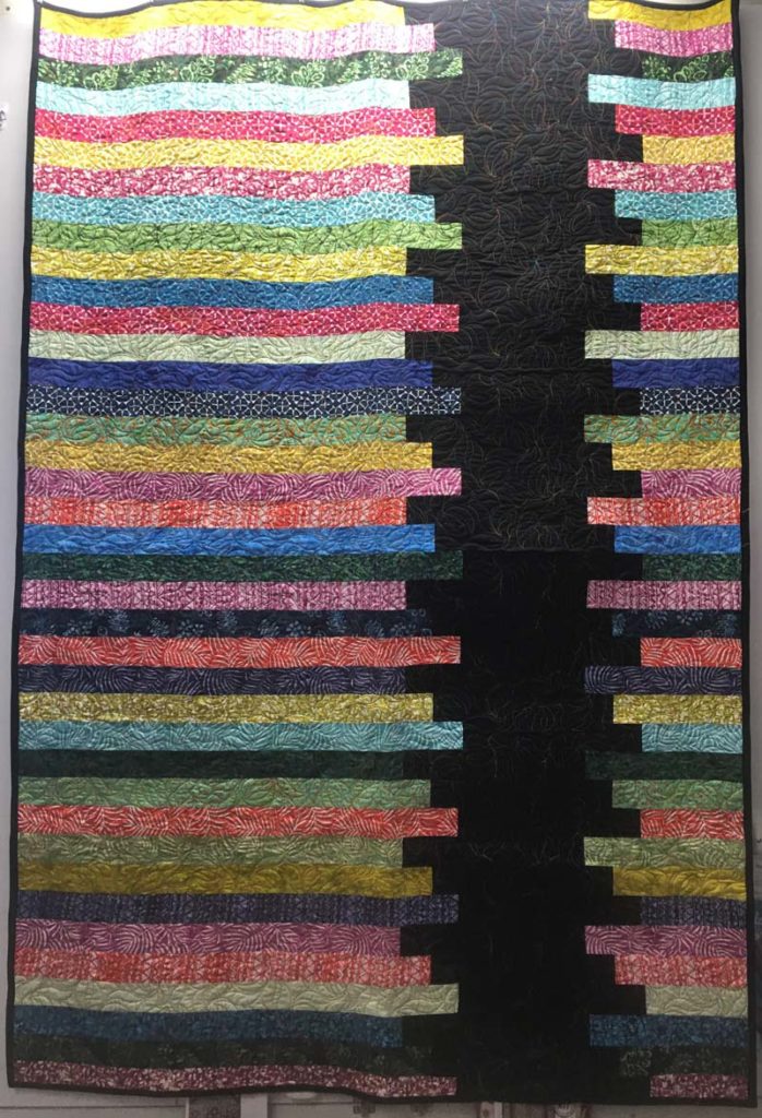 A large quilt made with a jelly roll (or more). It features a black jagged stripe on the left side going down, and horizontally many multi-colored stripes.