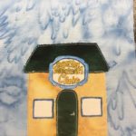 This quilt block shows a yellow square building with a trapezoid roof. The roof and the door are a dark green-blue. The door is in the center of the building with two white square windows on either side. The white windows are outlined with blue pen. The big draw to the block though is the belt-buckle like sign above the door. It's a huge blue bordered fancy sign with stylized yellow lettering that reads,