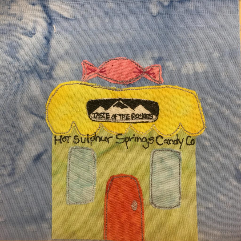 This quilt block shows a lime-yellow building with a bright yellow roof. The roof has a bottom trim like a candy wrapping, with rounded pieces. On the yellow roof is a giant pink twisted candy package, and below that on the front of the roof is black and white sign that has mountains on it and the words,