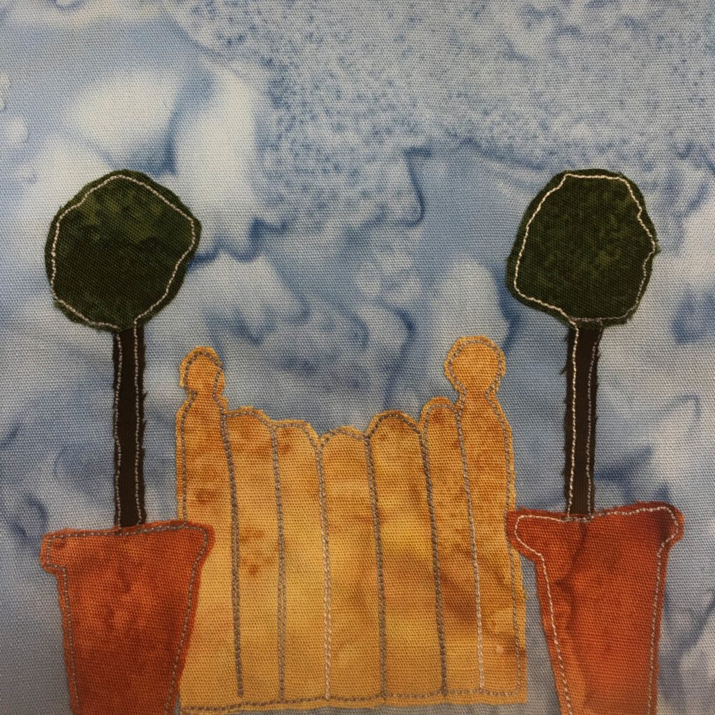 This quilt block shows a small wooden (orange-tan) gate with two topiary plants on either side. The topiary plants are in dark orange terracotta pots with long black stems/trunks, and a blob of dark green foliage at the top. These are outlined by a gray and white stitching. Each slat in the gate is also outlined by a gray and white stitching.