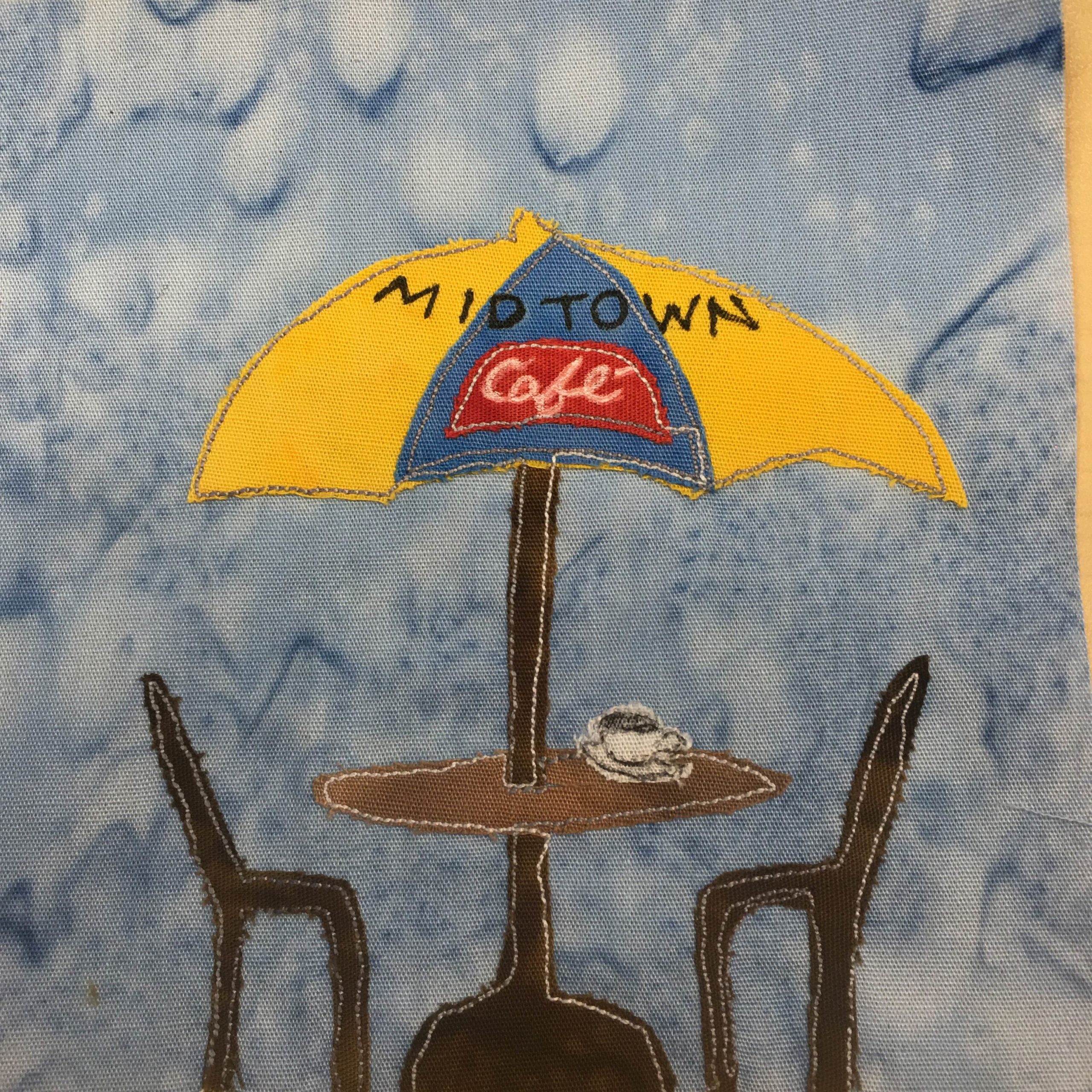 This quilt block shows a table with an umbrella over it and two chairs. The umbrella is coming out of the center of the table. The umbrella itself is a bright yellow and blue in sections. Across the top of the umbrella is the word, "Midtown" and underneath it in a red piece of fabric is written "Café" in white scripty pen. The umbrella pole and the chairs are a dark brown with light colored stitching outlining them. On the lighter brown table is a white piece of fabric shaped like a coffee mug with coffee in it.