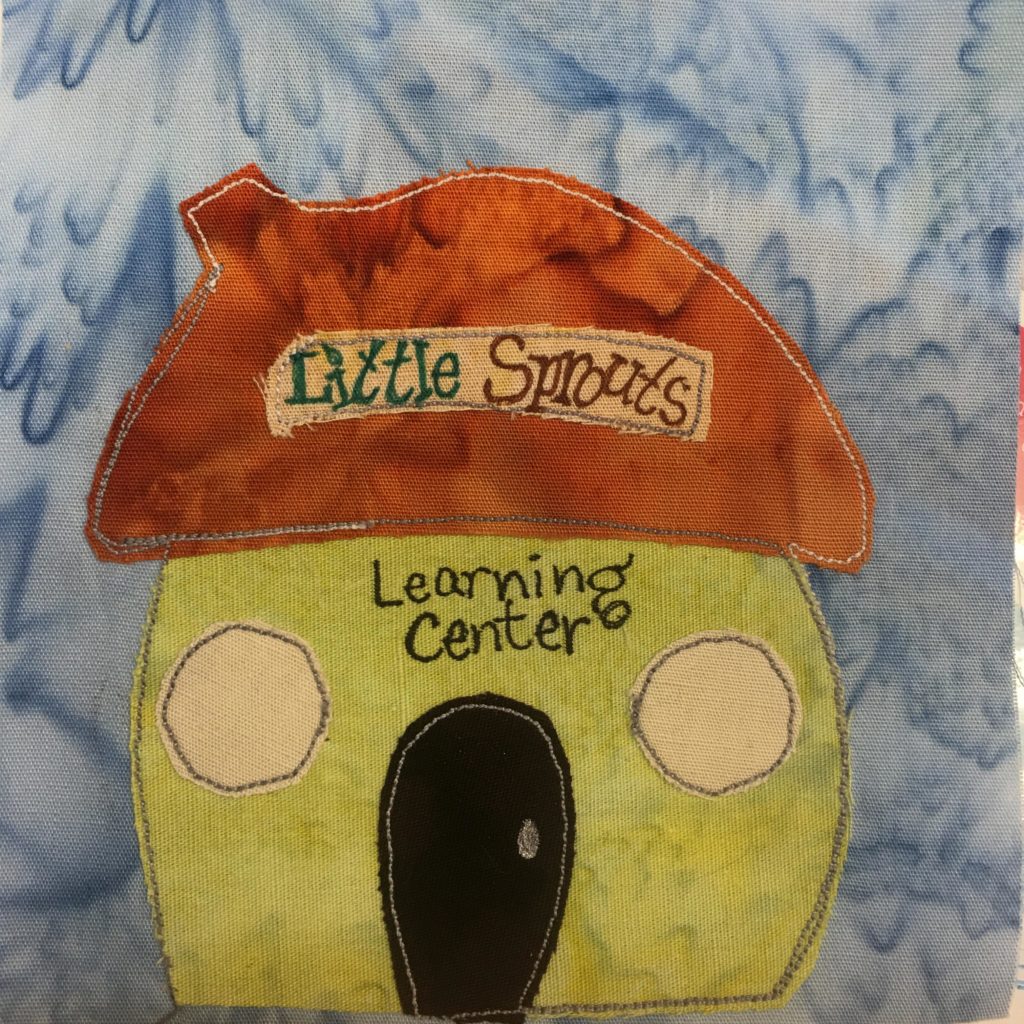 This quilt block shows a building that's made in the shape of an acorn, with a little wide stem coming out from the roof slightly on the left. The roof is a dark orange color with a tan sign on the front with the words,