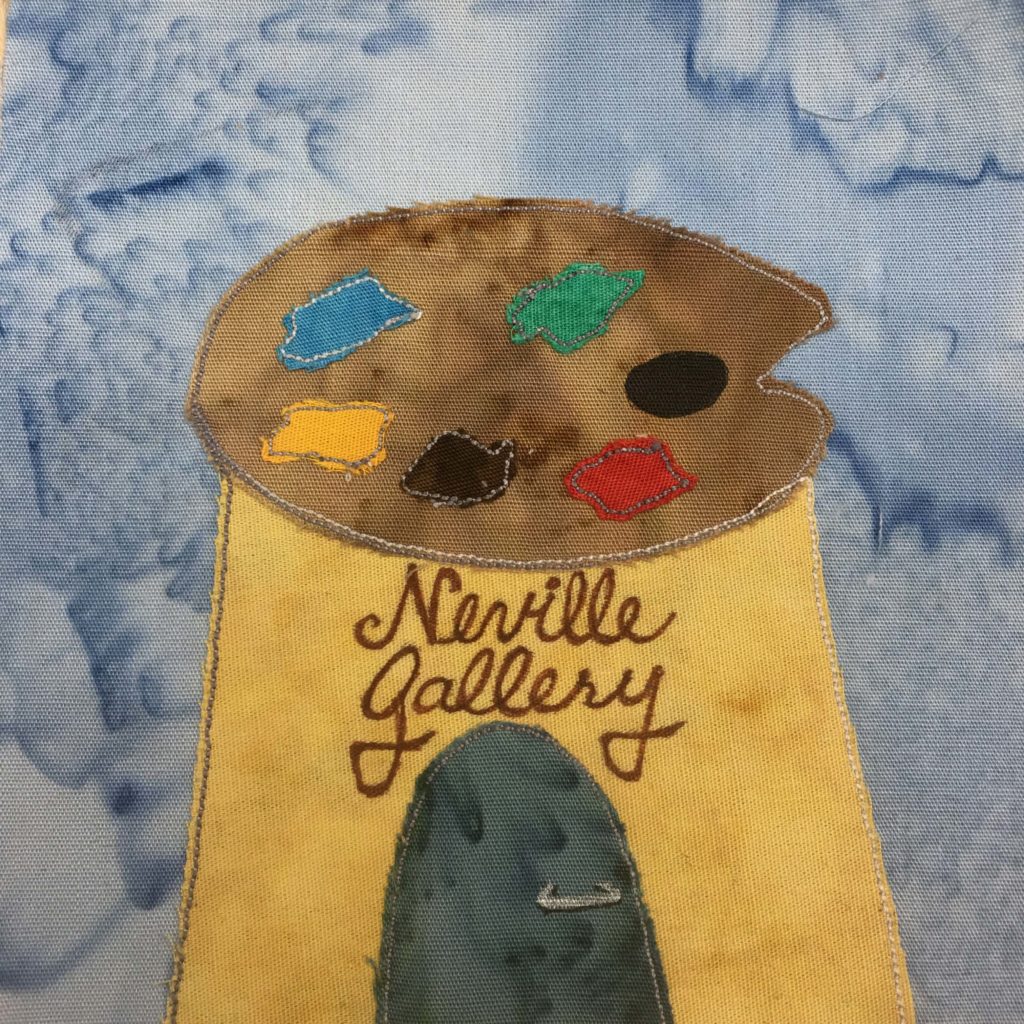 This quilt block shows a funny shaped building against a blue salmon-patterned background. The building itself is in a sort of trapezoid shape and is light yellow. The roof is actually a painter's palette, the base color being brown. On the palette are blobs of other color, including red, black, yellow, blue and green shown by accompanying fabrics. Below the giant painter's palette are the words in cursive, "Neville Gallery" written in brown on the yellow building. Below the words is a dark teal rounded door with a gray door handle.