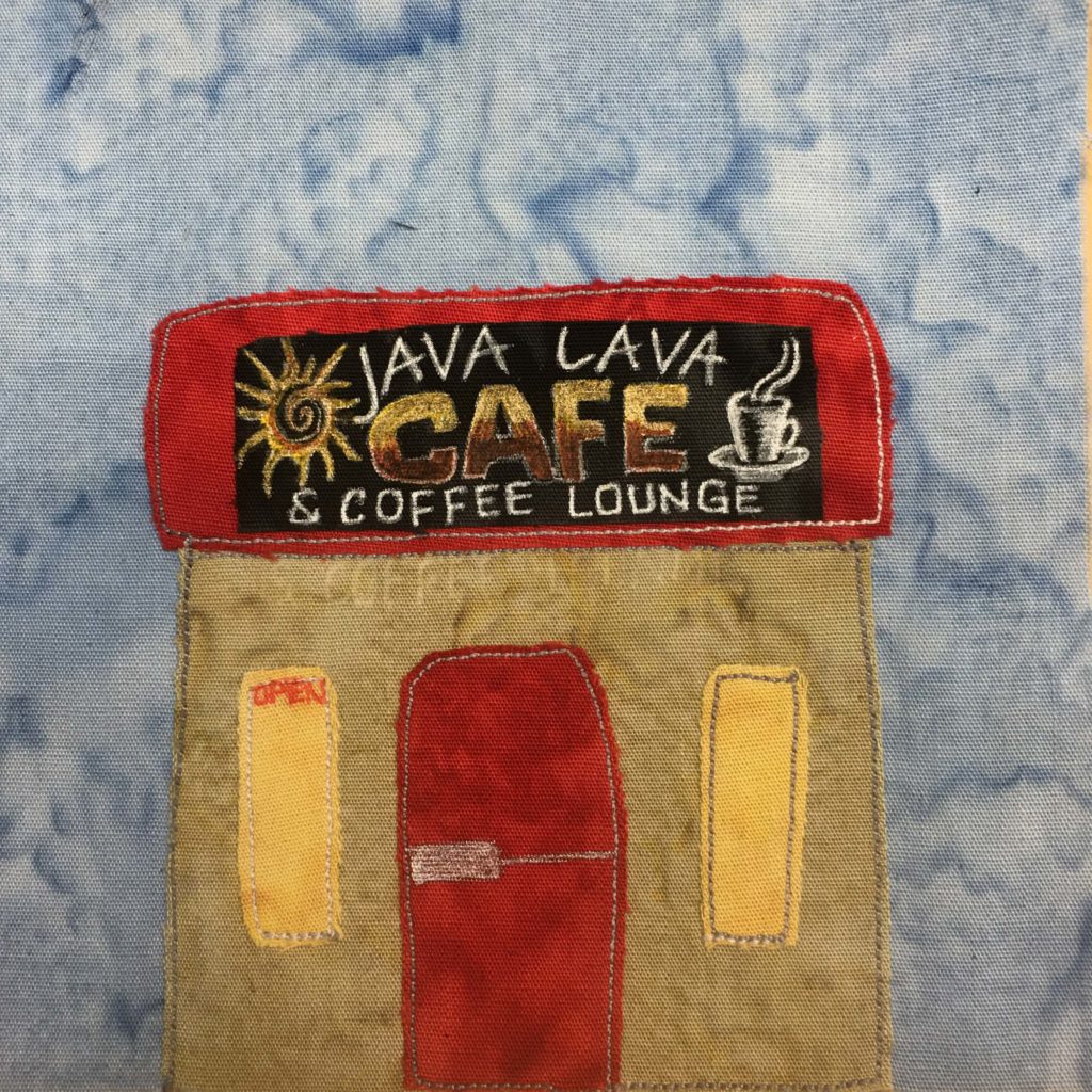 This quilt block shows a square building against a light blue salmon-patterned background. The building has a square flat roof that is a deep red. The door is also a deep red and pretty tall. On the red roof is a black sign with a lot of pen drawing on it. There is a sun with a spiral in the center, and a coffee cup with steam coming out o the top. The words, "Java Lava Cafe & Coffee Lounge" are on the sign in white, except for the word Cafe which is very large, center stage, and has a yellow-brown gradient. On either side the tall red door are tall yellow windows. In the left one there is the word "Open" in red letters.