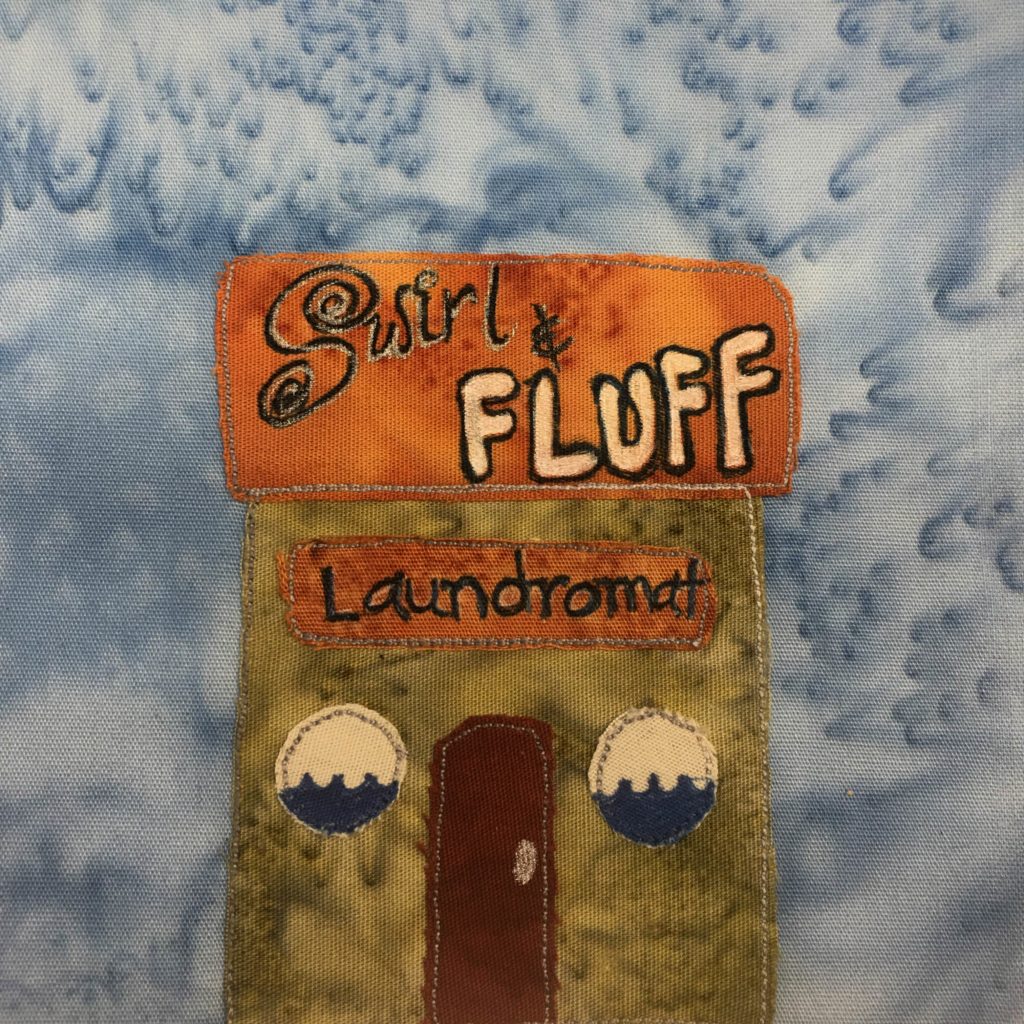 This quilt block shows a square building with a square flat roof. The roof is orange with the words, "Swirl & Fluff" written on it. The swirl is in black and white pen, and the S has swirls on it. The fluff is in bold white with black pen outlines. Below the orange roof is a green building with a darker orange sign on it that reads, "Laundromat." The windows on either side of the red door are round. They each have waves of water on the bottom half, as if one is looking into a washing machine. The door is red and in the center.