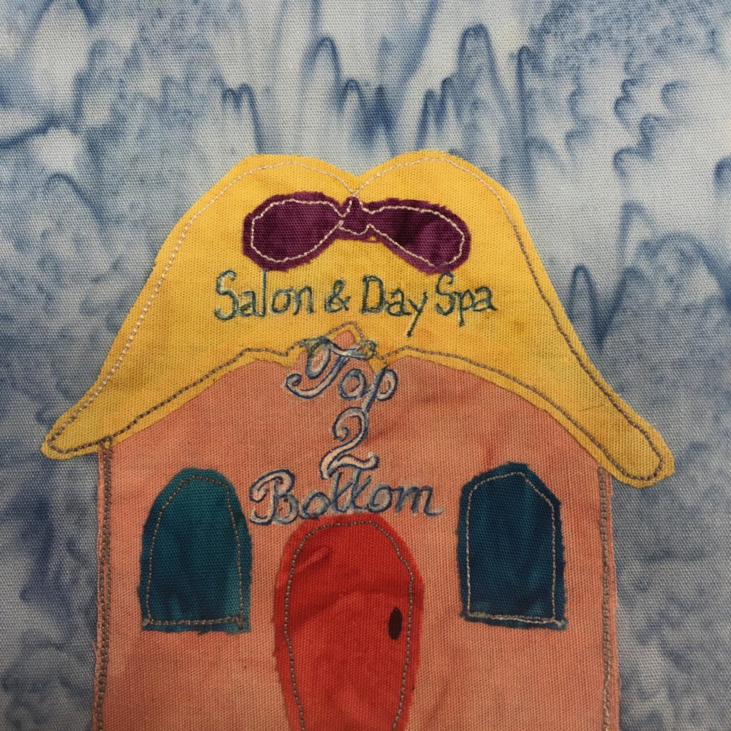 This quilt block shows a square building but with an outlandish roof. The roof looks like hair sitting on someone's head with a purple bow in it. The hair is yellow itself. On the hair and the building are the words, 