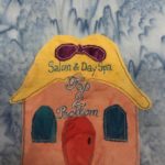This quilt block shows a square building but with an outlandish roof. The roof looks like hair sitting on someone's head with a purple bow in it. The hair is yellow itself. On the hair and the building are the words,