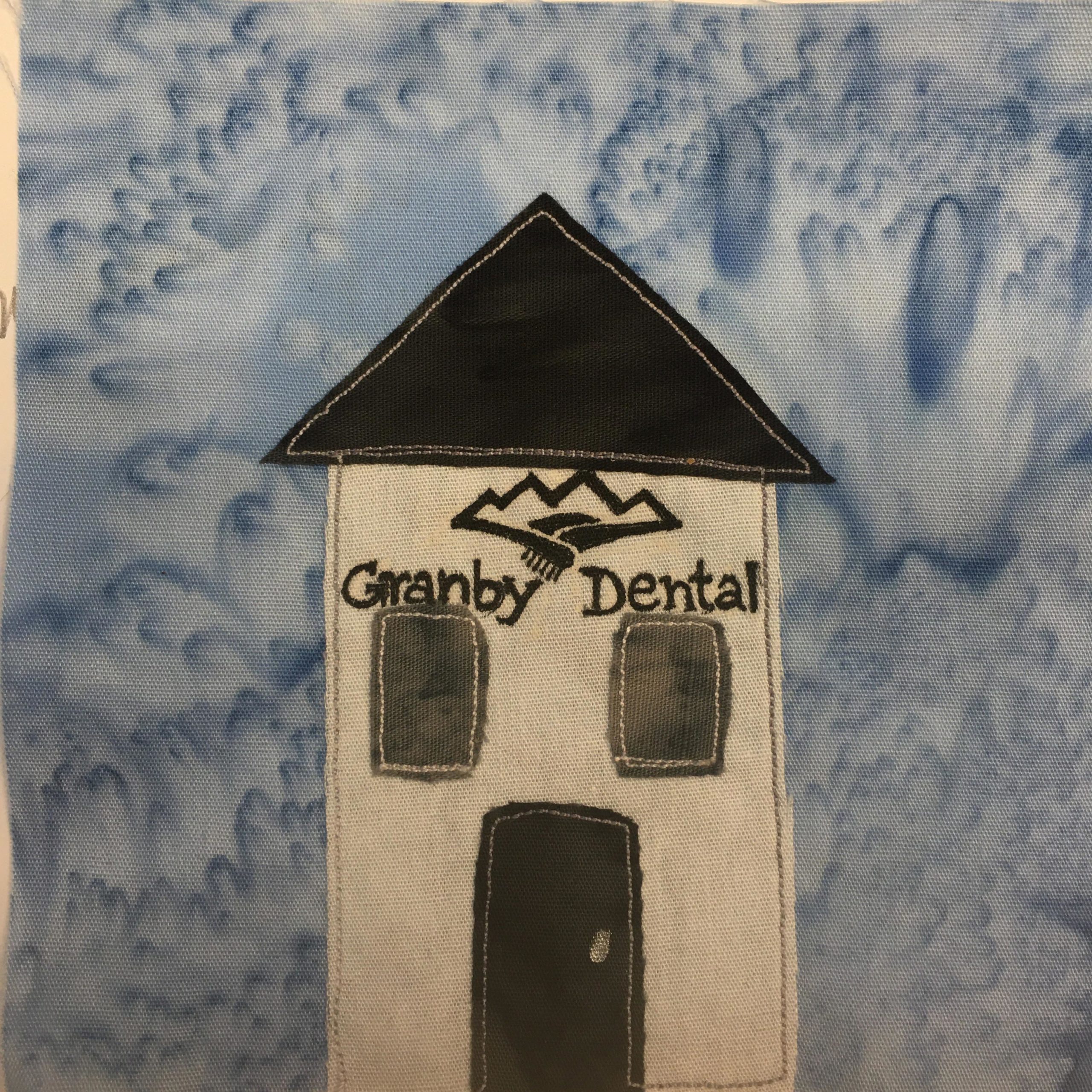 This quilt block shows an austere building against a light blue salmon-patterned background. The building is white and tall with a very triangular black roof. There are two dark grey second story windows outlined with white stitching. The door is an even darker shade of gray. The building and roof itself are outlined in gray stitching. Above the two second-story windows is writing that says,