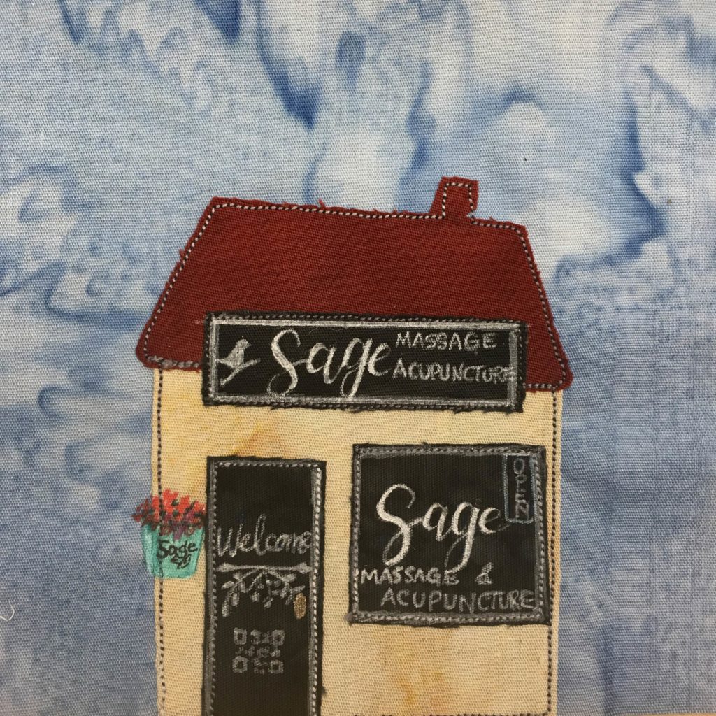 This quilt block shows a shop building. The roof is a deep matte red with a little chimney poking out of the top right. It's in a trapezoid shape and is slightly tilted to the right. There is a black sign on the separation between the building and the roof that reads,